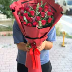 Mat kırmızı uzun ambalajda kırmızı gül, cipso ve okaliptus buketi; kurdele üzerinde tek gül