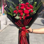 Siyah ambalajın içinden kırmızı pliseler görünen kırmızı gül buketi, organze kurdele ve tek gül